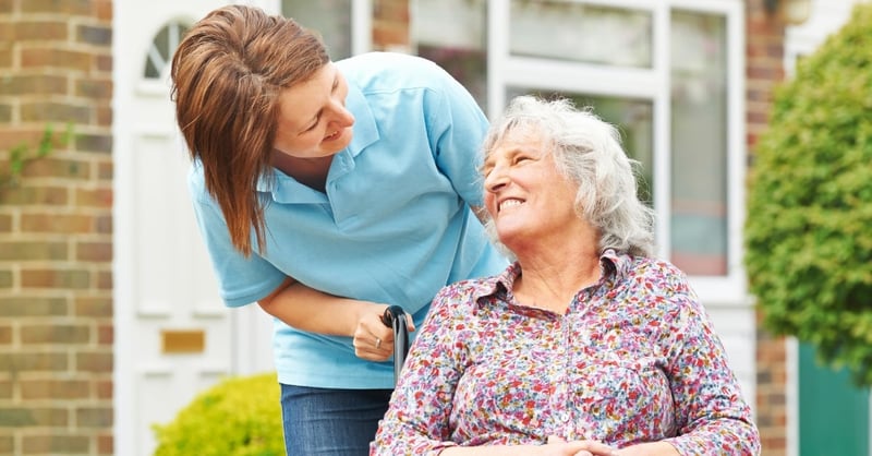 A smiling elderly lady in a wheelchair being pushed by a care worker
