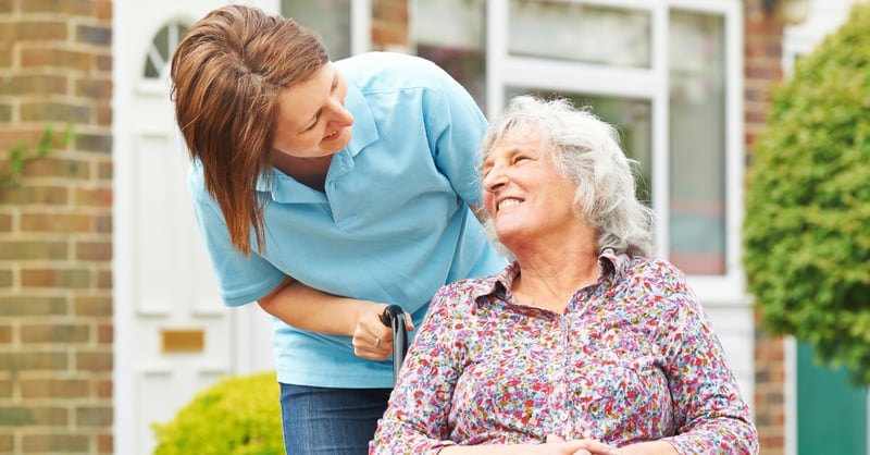 A smiling elderly lady in a wheelchair being pushed by a care worker