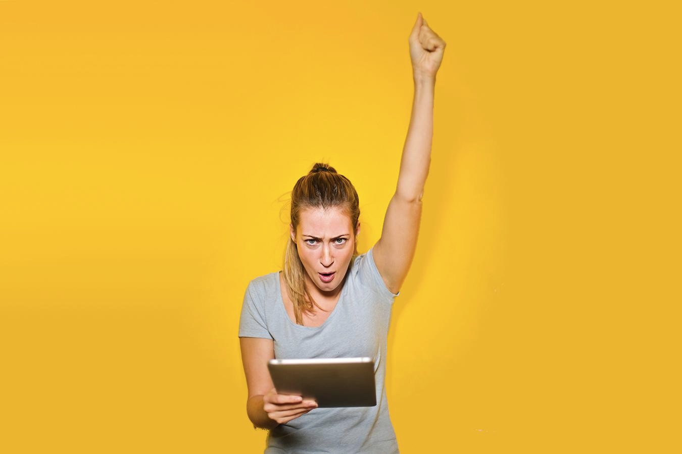 A young woman holding a tablet and punching the air, against a bright yellow background