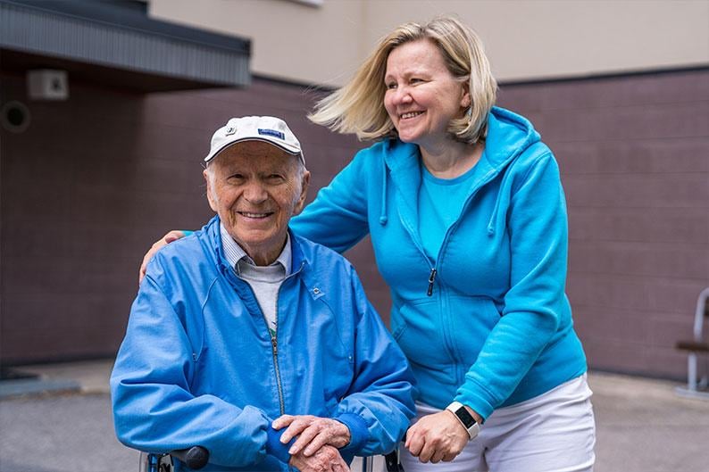 A female carer with her arm around an elderly male man in a white cap, both wearing blue hoodies