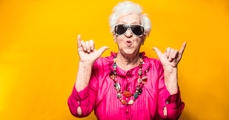 A lively looking elderly lady in sunglasses dressed in pink clothes against a bright yellow background