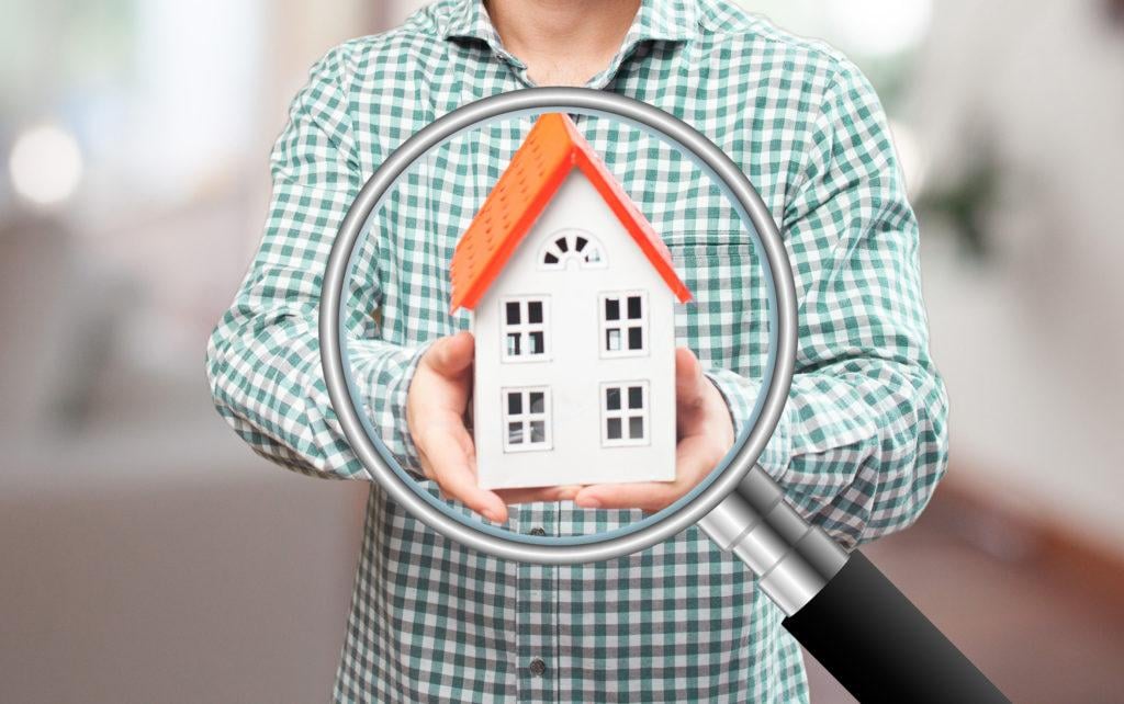 A person in a checked shirt holding up a model of a house with an orange roof behind a magnifying glass